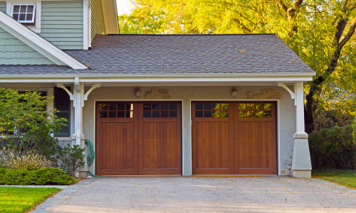 Image of a residential house with a two-car garage featuring wooden doors. The garage is framed by white trim and surrounded by greenery, with sunlight filtering through the trees in the background.